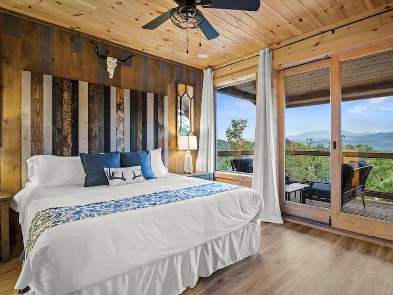Bedroom of Pigeon Forge rental, with rustic style detailing, and French door access to a deck with outdoor seating and views of forested mountains.