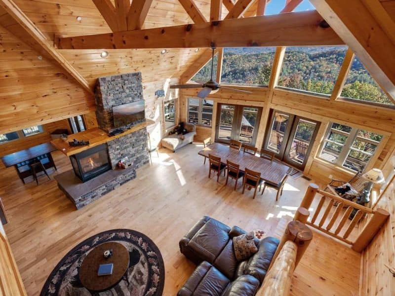 Living area of Pigeon Forge vacation cabin, with vaulted ceilings with exposed beams, wood paneling throughout, stone fireplace, and floor to ceiling windows looking out at forest and mountain views.