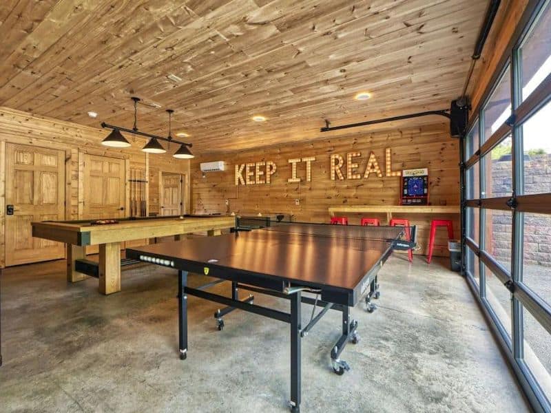 Game room of Pigeon Forge Cabin, with pool table and ping pong table, with wood paneling, industrial details and large garage door opening to the exterior.