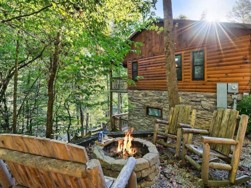 Outdoor fire pit adjacent to cabin, with stone and wooden accents. surrounded by trees.