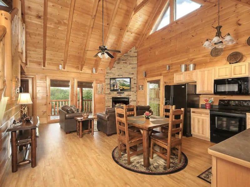 Living area and kitchen of Pigeon Forge vacation. cabin, with foloor to ceiling wood paneling, pitched roof with exposed beams, seating and dining area, with a fireplace and access to balcony.
