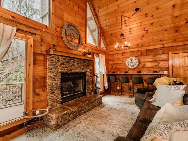 Living area of Pigeon Forge vacation home, with stone fireplace, modern decor, floor to ceiling wood paneling, and large windows with views of forest and creek.