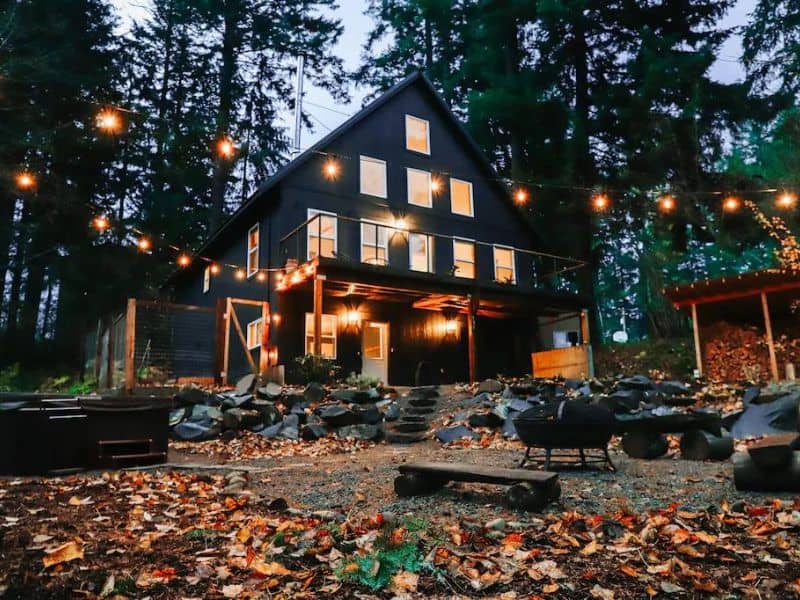Black cabin lit up at dusk with lots of windows and an outdoor fire pit with string lights. Part of a guide to the best cabin rentals near Mt Rainier