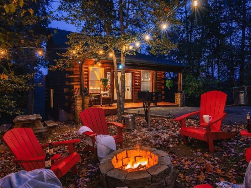 Small log cabin at night lit up with fairy lights with an outdoor fire pit and red Adirondack chairs