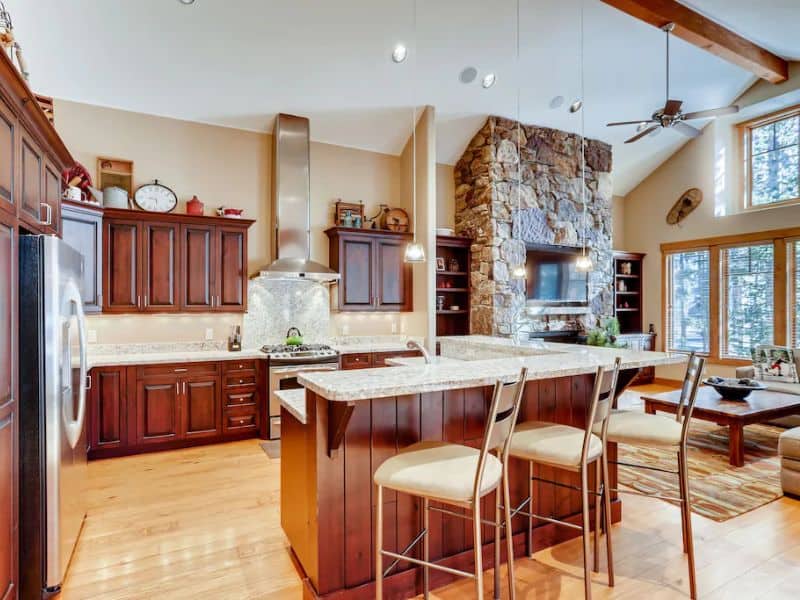 A large kitchen in with wood cabinets and stone counter tops with a view of a large stone fireplace in the living area of a ski in ski out vrbo in Breckenridge, CO.
