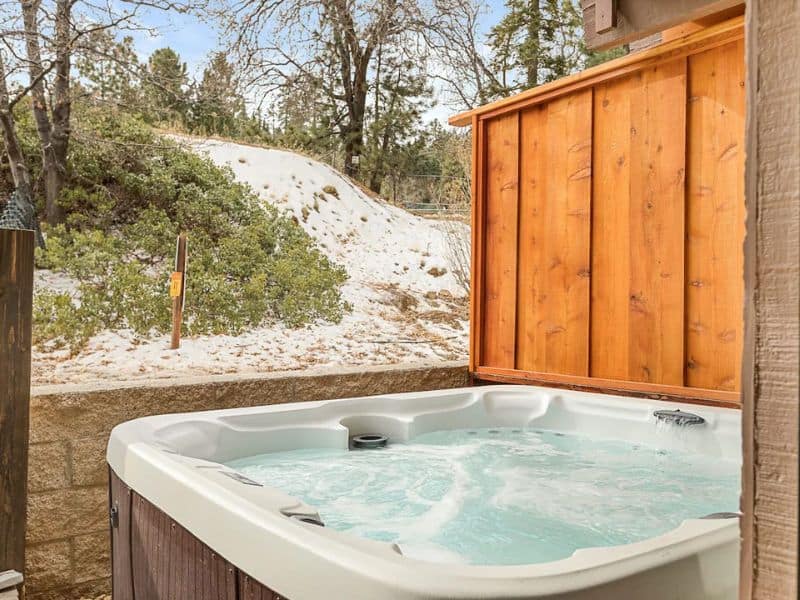 Hot tub on the deck of a Big Bear winter cabin with a wooden privacy screen and a snowy hillside in the background.