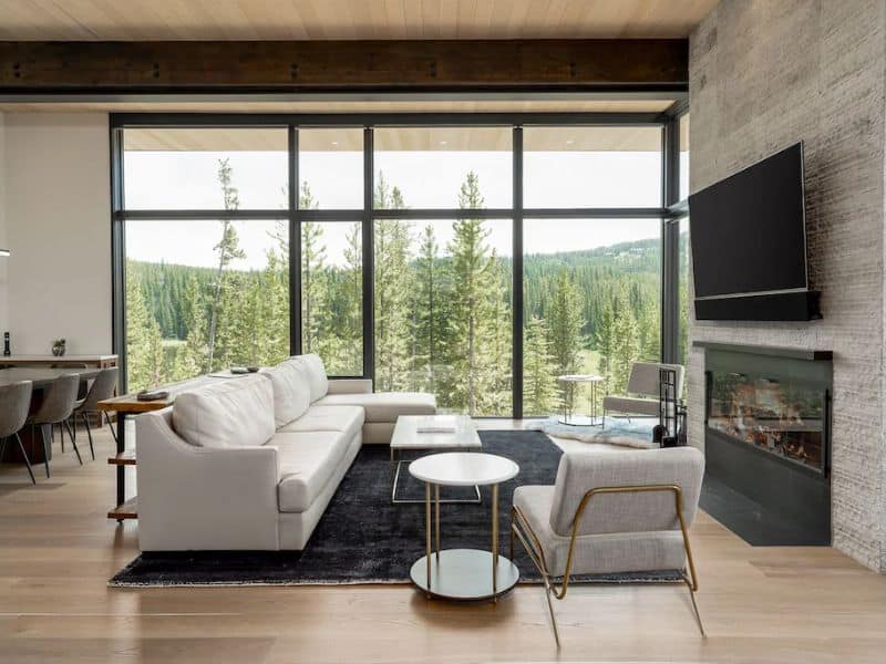 Spacious living area with floor-to-ceiling windows offering forest views, a fireplace, and contemporary furnishings in a luxurious ski in ski out Big Sky Montana rental home.