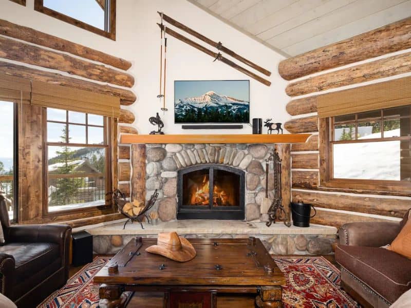 Elegant and rustic living room inside a ski in ski out Big Sky Montana rental, featuring a stone fireplace with a warm fire and a mounted flat-screen TV, surrounded by log walls and large windows with a snowy view.