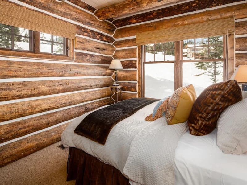 Cozy bedroom with log walls, plush bedding, and a window view of snow-covered trees in a Big Sky Montana ski in ski out cabin rental.
