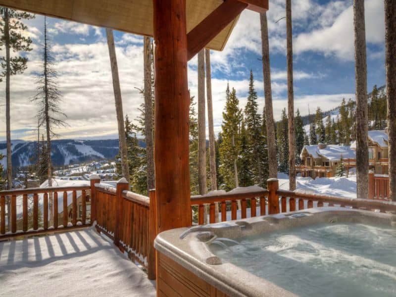 Hot tub on a wooden deck with a stunning view of snow-laden trees and mountain vistas, a highlight of this ski in ski out Big Sky Vrbo rental.