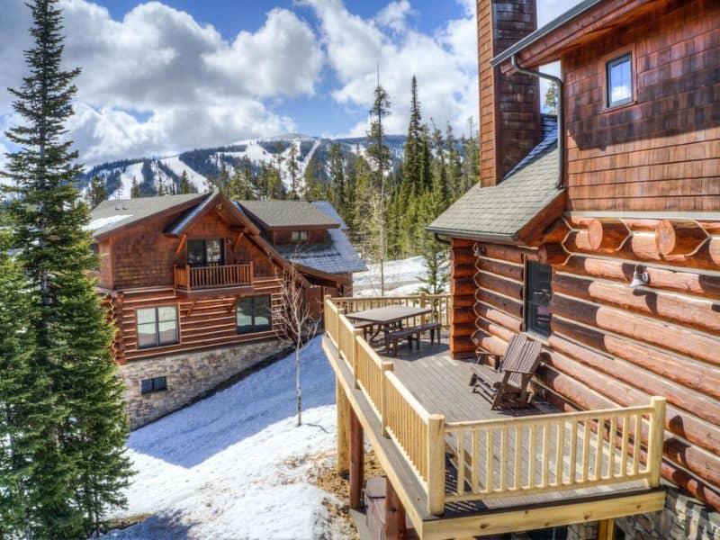 Exterior view of a log cabin with a deck: one of the best ski in ski out Big Sky MT rentals, with mountain slopes in the background.