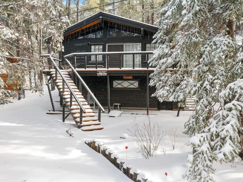 Snowy exterior of a modern black Big Bear winter cabin with a raised deck, showing a contrast against the white snow and green pine trees.