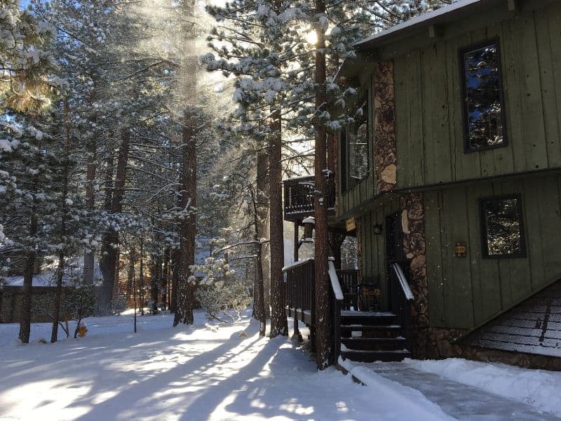 Snow-covered grounds with sunlight filtering through the pines at a Big Bear winter Vrbo cabin, featuring a green exterior.