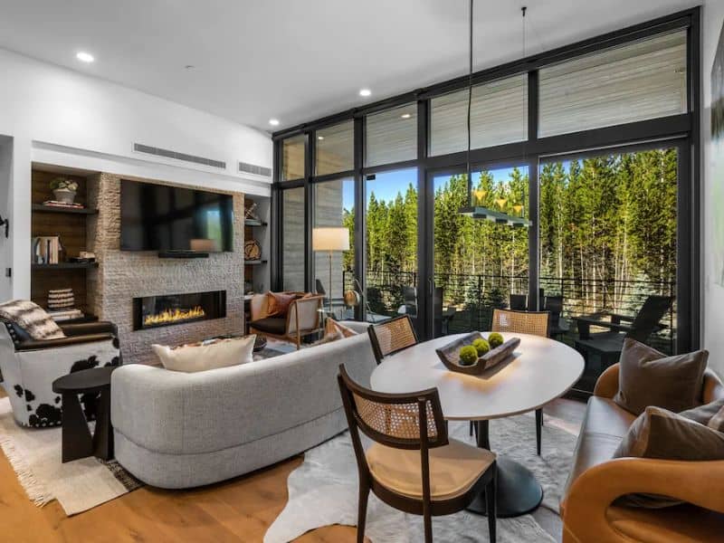 A contemporary living room inside a ski in ski out Big Sky Montana rental, with plush seating, a stone fireplace, and large windows revealing the forest outside.