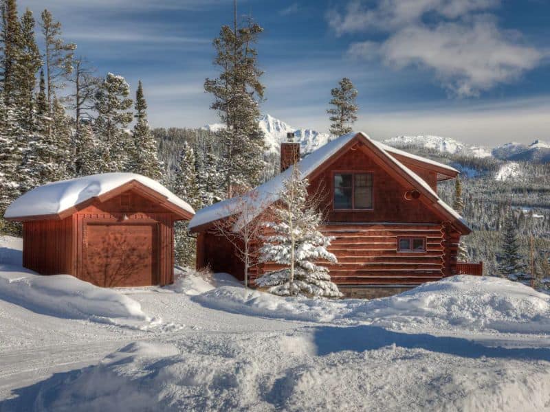 Log cabin rental with a garage surrounded by snow-covered trees in a Big Sky MT, with mountain views in the distance.
