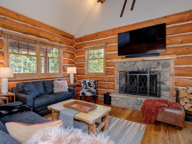Living room with a stone fireplace, large windows, and wooden beams in a cabin rental in Big Sky Montana.
