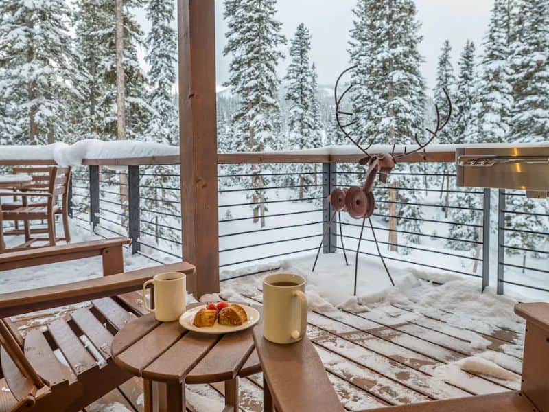 Balcony with a snow-covered railing and forest view from a ski in ski out Big Sky Vrbo cabin, with breakfast set on the table.