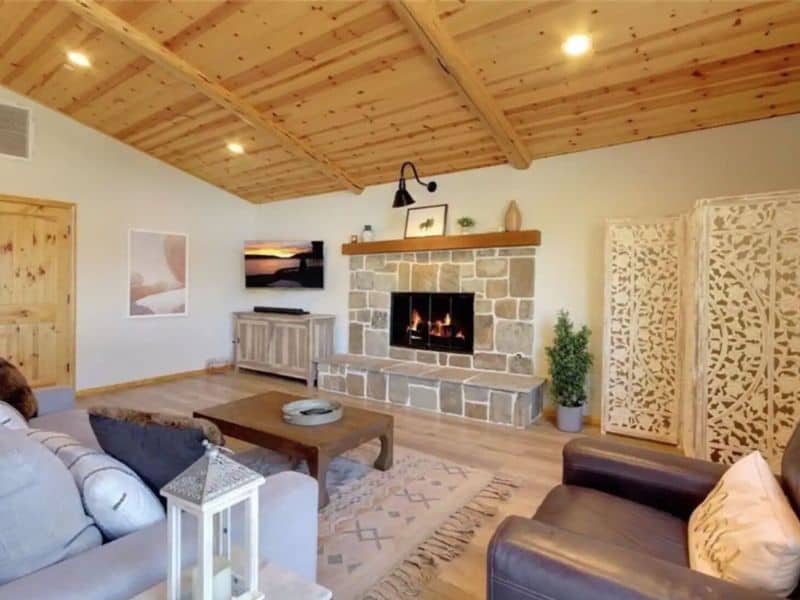 Spacious living room inside a cabin rental near Bear Mountain in Big Bear, with vaulted wooden ceilings, a stone fireplace, and comfortable seating arranged around a rustic coffee table.