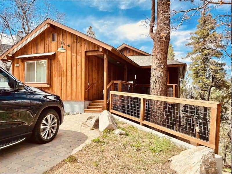 Front view of a cozy Big Bear winter cabin rental with a wooden façade, featuring a wraparound deck and a parked SUV, set against a backdrop of mature trees.