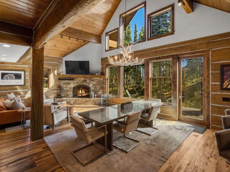 Large open-plan dining area with a stone fireplace and large windows looking out to the forest in a Big Sky Montana Vrbo rental.