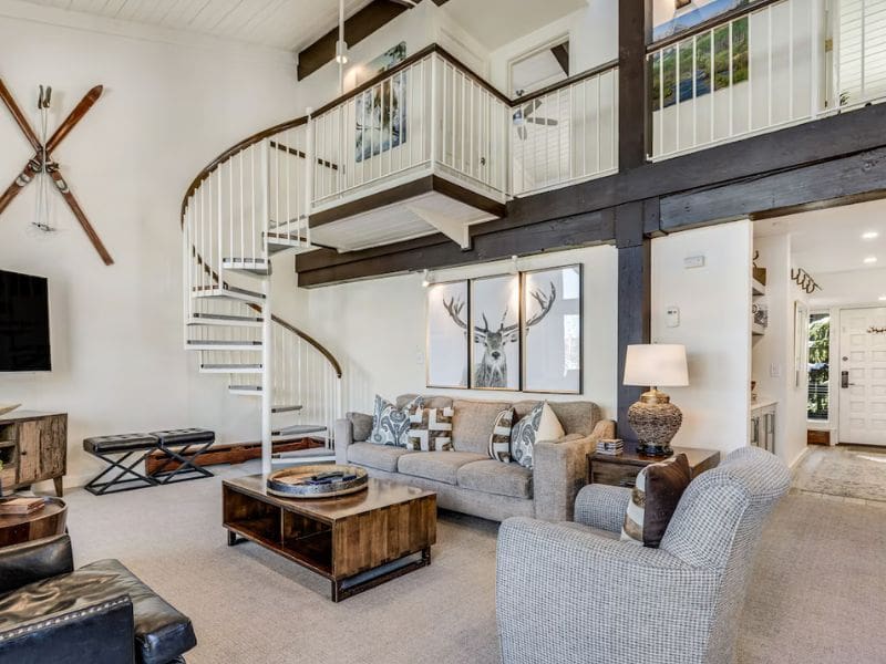 Stylish living room in an Aspen Ski In Ski Out rental with a modern, open design featuring a spiral staircase leading to an upper loft. The room is furnished with a gray sofa, wooden coffee table, and ski-themed decor, including mounted vintage skis on the wall. There are large windows and a high ceiling.