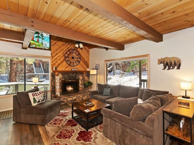 Cozy living area inside a cabin near Snow Summit, featuring a stone fireplace, plush seating, and a large window with a forest view.