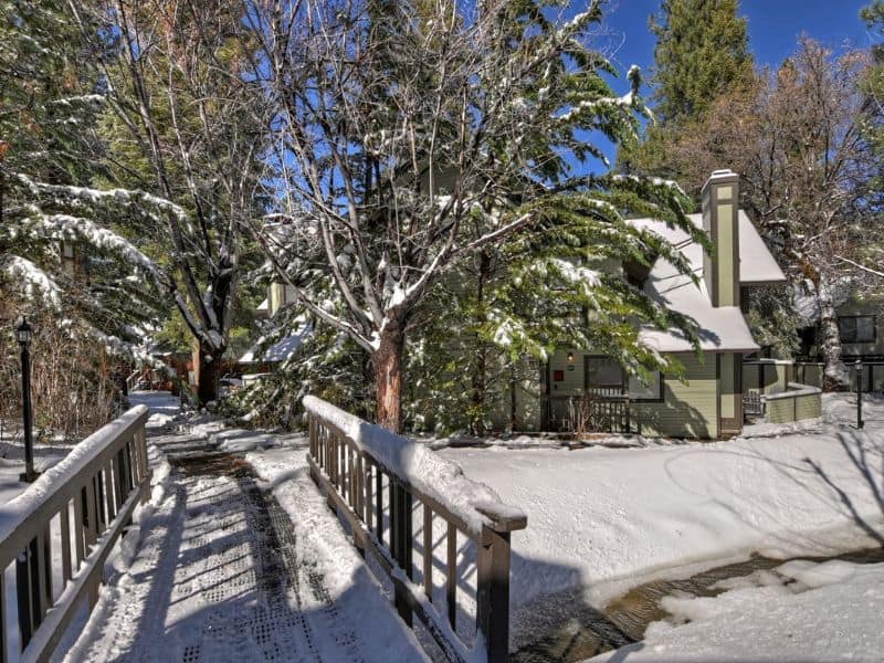 Snow-covered path leading to a modern Big Bear winter Vrbo rental, nestled among tall pines with a clear blue sky above.