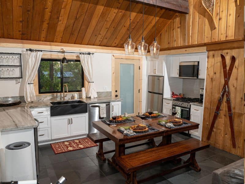 Rustic Big Bear winter cabin dining area with a wooden table set for a meal, flanked by vintage ski equipment and a classic white kitchen in the background.