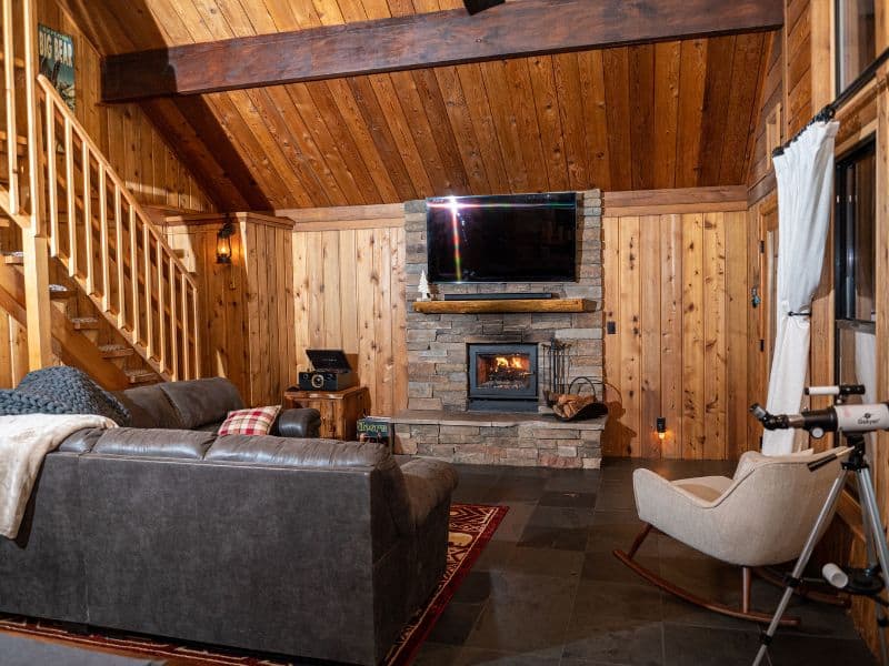 Cozy interior of a cabin rental near Bear Mountain in Big Bear featuring a plush sofa, rustic stone fireplace, and a warm wooden staircase leading to an upper level.