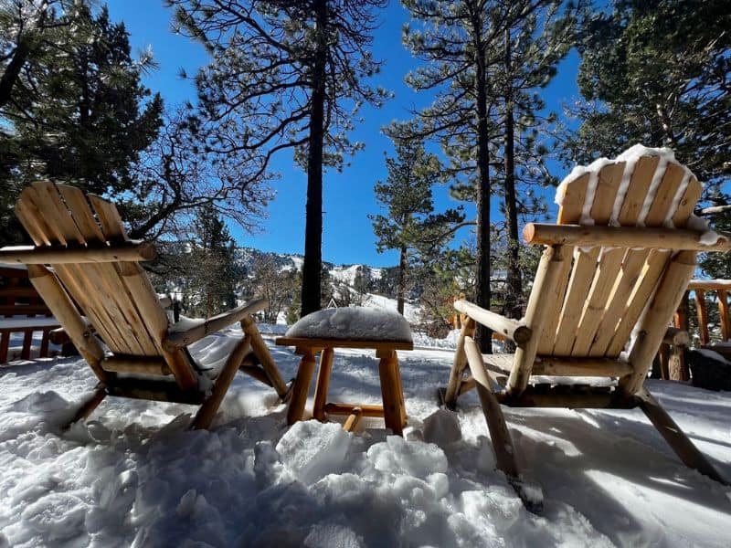 Two Adirondack chairs covered in fresh snow, facing a view of tall pine trees and ski slopes in the distance in Big Bear CA.