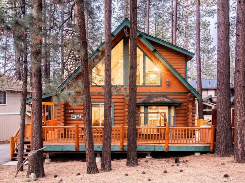 Log cabin rental in Big Bear with large windows and a wraparound deck nestled among pine trees.
