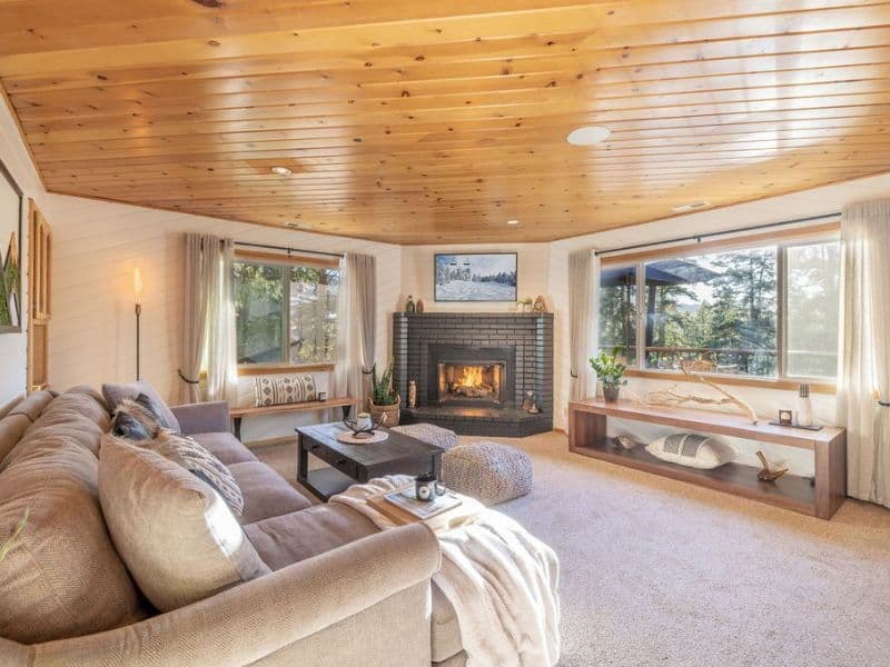 Cozy living room with a fireplace in a cabin rental near Bear Mountain in Big Bear, complete with comfortable sofas and large windows offering forest views