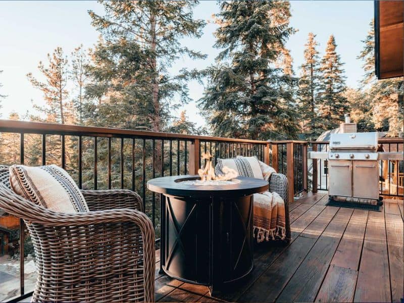 Spacious outdoor deck of a Big Bear winter cabin rental, featuring wicker chairs and a fire pit table, with a barbecue grill and a forested backdrop.