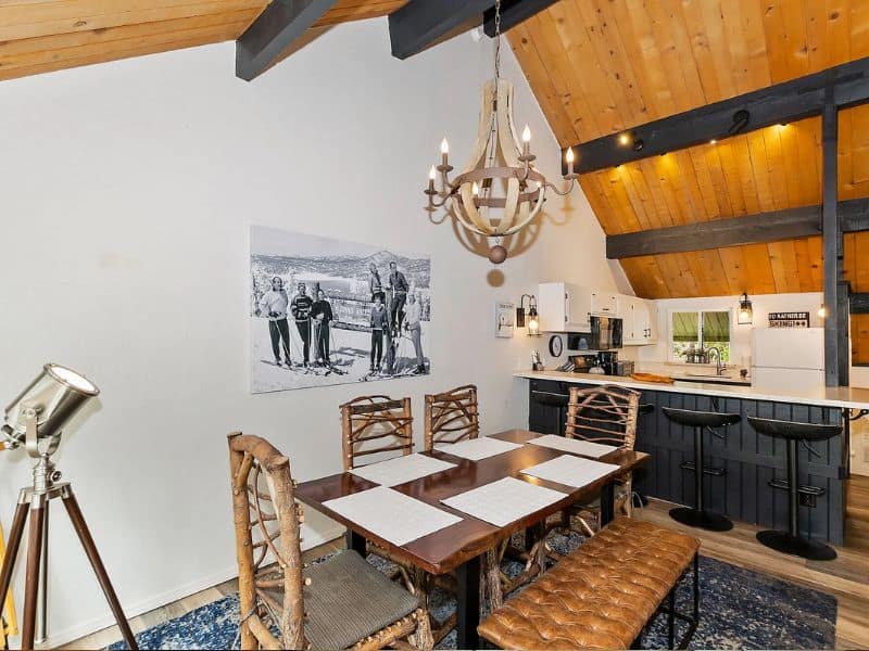 Dining space with a rustic chandelier and a full kitchen in the background. Cabin near Snow Summit in Big Bear.