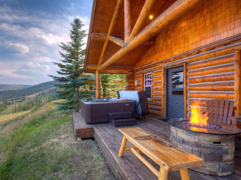 Relaxing deck of a log cabin with a hot tub and fire pit overlooking the mountains at a ski in ski out Big Sky MT rental on Vrbo.