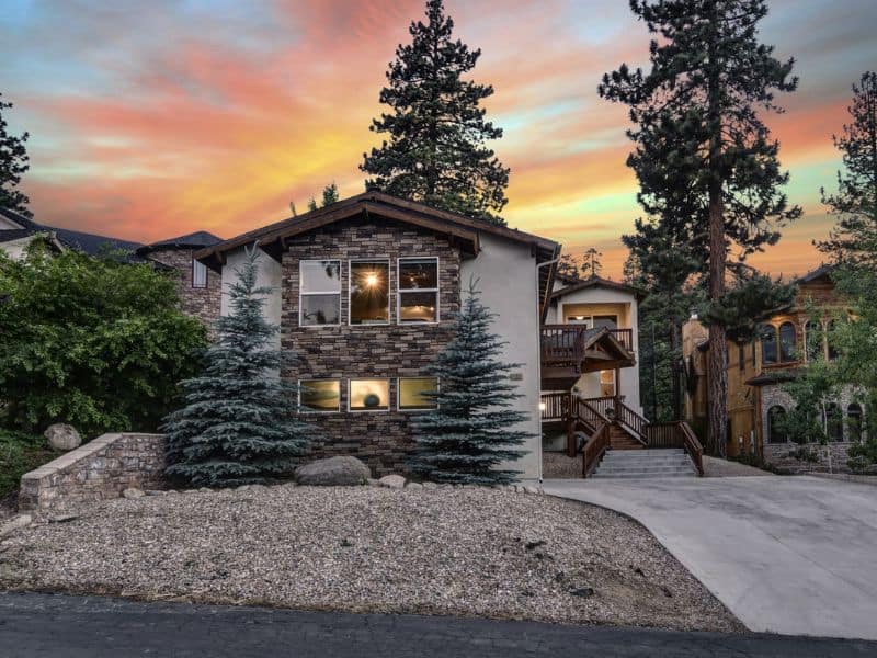 Twilight sky with hues of orange and purple as a backdrop to a modern Big Bear vacation home with stone accents and tall pine trees.