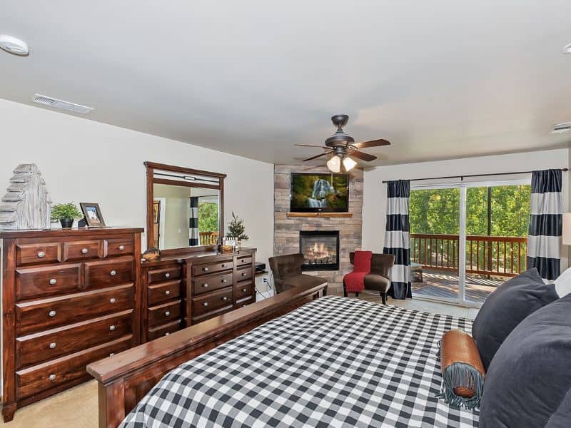 Bedroom in a cabin rental near Bear Mountain in Big Bear featuring a large checker-patterned bed, stone fireplace, and balcony access with forest views.