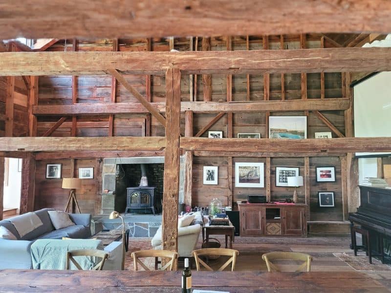 Interior of a cozy Catskills cabin rental with exposed wooden beams and a lofted ceiling, a comfortable living area with a couch, chairs, and a warm fireplace.