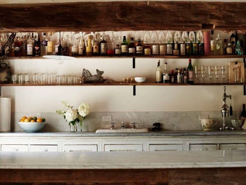 Well-stocked open shelving in a Catskills rental cabin, displaying an array of glassware and a diverse selection of spirits and liqueurs above a white countertop.