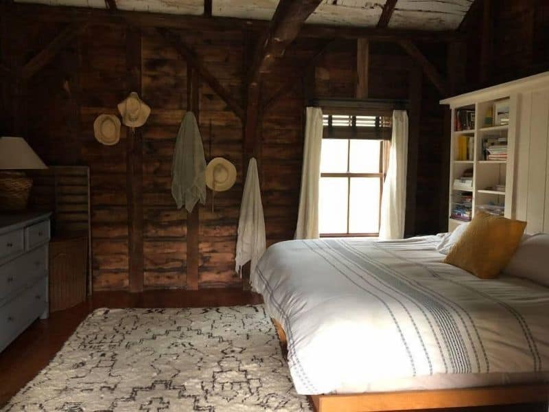 A rustic bedroom in a Catskills cabin rental with a plush bed with white linens, rustic wooden walls, and hats and towels hung on the wall.
