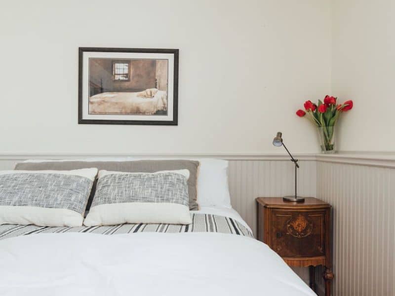 Charming bedroom in a cozy cabin in the Catskills, featuring a comfortable bed with gray-striped bedding, a vintage nightstand, and fresh tulips adding a pop of color.