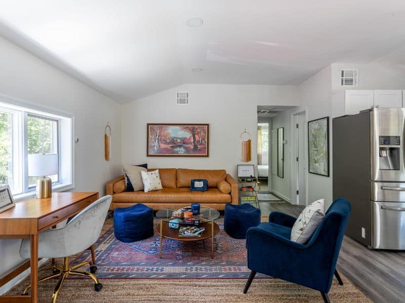 Bright and airy living area in a Catskills rental featuring a camel-colored leather couch, two blue velvet chairs, and a workspace with a vintage-style desk.