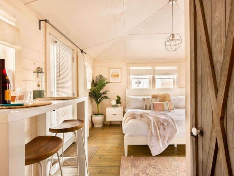 Cozy interior of a small Catskills rental cabin with a kitchenette and breakfast bar leading into a sunlit bedroom area with plush bedding and soft textures.