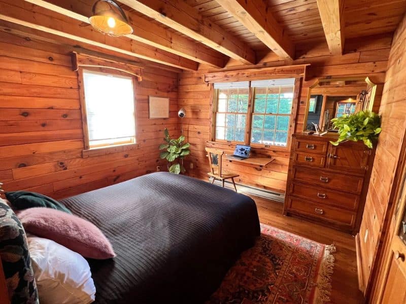 Warm and inviting bedroom in a winter Catskills cabin rental, with natural wood walls, a large bed with dark bedding, and a sunny nook with a desk.