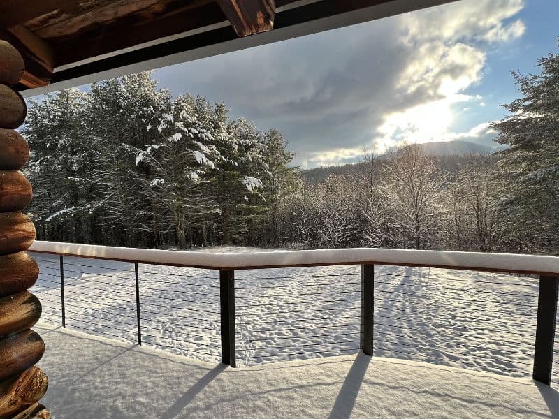 Snow-covered balcony view from a cozy cabin in the Catskills, with a backdrop of snow-dusted trees and a clearing sky.