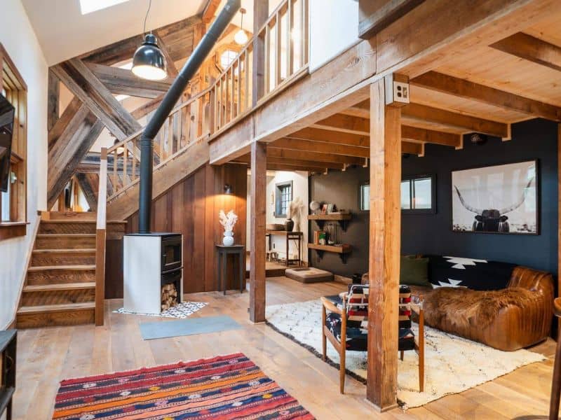 Open living space inside a cozy Catskills cabin rental with a modern wood stove, staircase leading to a loft, and eclectic furnishings against a rustic backdrop.