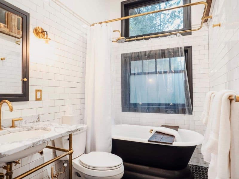 Chic bathroom in a winter Catskills cabin rental, with white subway tiles, gold fixtures, a freestanding black bathtub, and a classic pedestal sink.