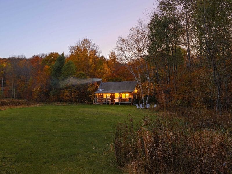 Evening view of a secluded Catskills cabin rental with glowing windows, set against a backdrop of autumn-colored trees.