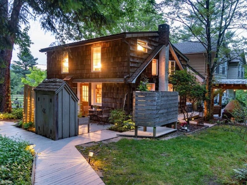 Twilight scene of a cozy Catskills cabin rental with warm interior lights, surrounded by greenery.