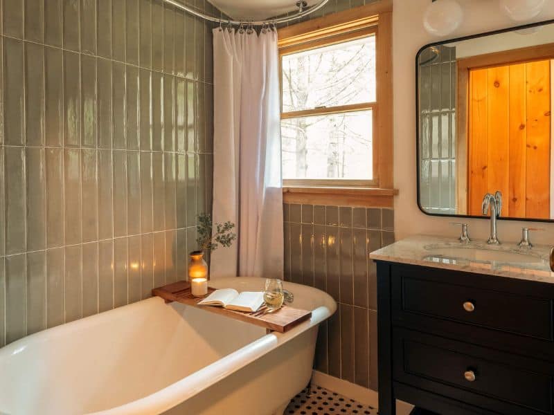 Charming bathroom in a cozy Catskills cabin rental, featuring a clawfoot tub with a wooden bath tray, subway tiles, and a view into the forest.